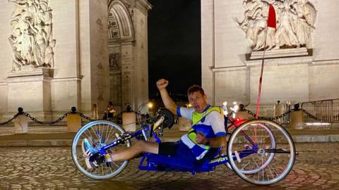 A man in an adapted bicycle. He is at a marble arch.