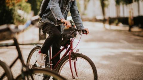 A stock image of a man wearing a denim jacket riding a purple bicycle on a street.