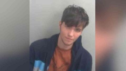 Chase Wiseman has short brown hair with a fringe in his police custody photo. He has a blue jacket and a camel brown T-shirt on.