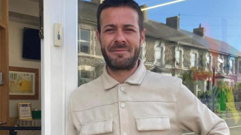 Luke Davies leaning against a large window. He is bearded with short dark hair and wearing a light coloured jacket 