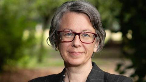 A woman with grey bobbed hair and black glasses wearing a dark blazer. She is outdoors with greenery behind her and smiling.