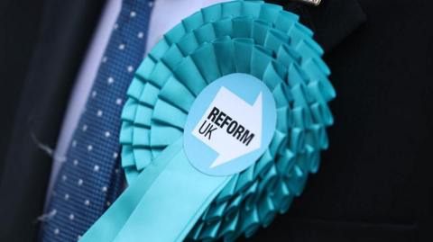 A light blue rosette pinned on a navy blue suit. The centre of the rosette has the Reform UK logo on it.