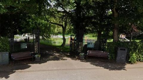 The entrance to Hall garth Park in Hornsea. Two benches are sited at the entrance, both in the shade of large trees. Slides and swings are behind the trees.