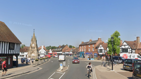 A street with shops, cars, and a clock tower in the centre, with people walking and cycling.