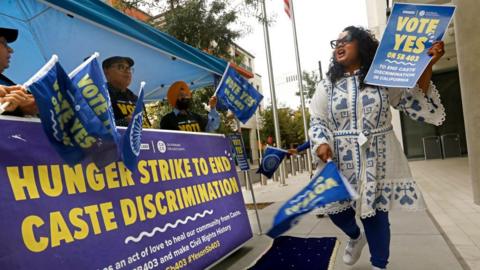 Woman in blue and white dress holding sign that says "Vote yes"in front of blue and yellow banner announcing hunger strike.