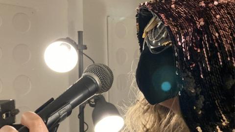 A female artists stands holding a microphone to her face which is covered by a black mask and she's wearing a sequinned headscarf covering her hair.