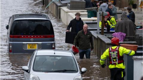 People wade through water on a street in Pontypridd, Wales, with an emergency worker in the foreground and two vehicles also partially submerged.