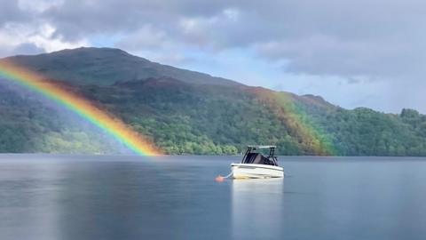 A rainbow appears to arc cross and end in the water in front of a mountain. A second sliver of rainbow sits a little further across to the left. In the foreground a small boat is attached to a buoy.