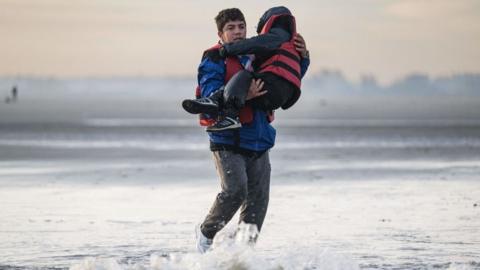 A man carries a child through the surf on a beach in northern France