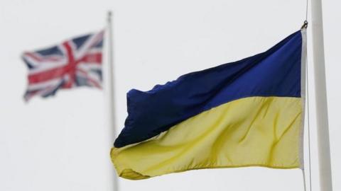 A Ukrainian flag is seen flying with a Union Jack in the background