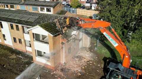 Two-storey brick-built flats being demolished by an orange excavator