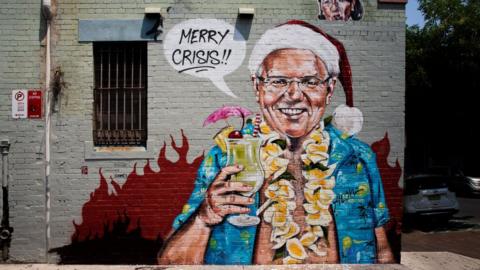 A mural of Scott Morrison depicted with Hawaiian motifs and a Christmas hat, saying in a speech bubble: "Merry crisis!!"