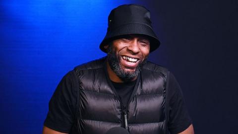 Flowdan, a black man, smiling at the camera. He's wearing a black bucket hat, tshirt and puffer jacket. The background is moody blue lighting.