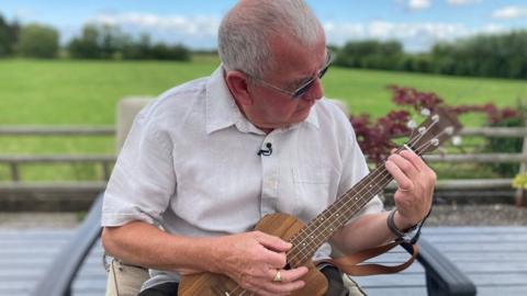 A man with white hair in a white shirt holding a ukulele