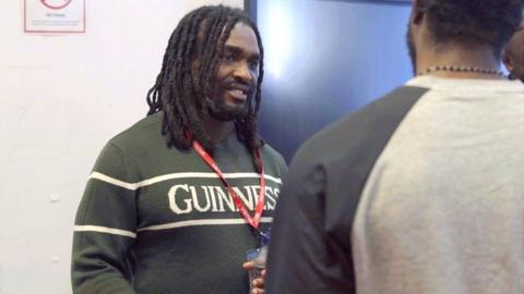 A man with dreadlocks wearing a green jumper with Guinness written across the chest smiles as he listens intently to a man in a grey sweater with his back to the viewer.