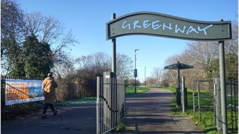 Green wave-shaped sign for the Greenway, with a concrete path, grass and trees either side