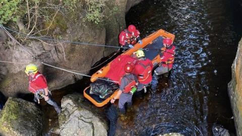 A man lies on a floating stretcher in water as volunteers in red jackets and helmets set up ropes to winch him out of a gully