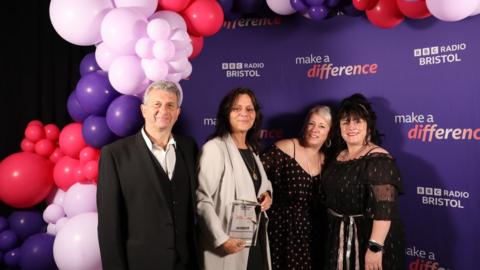 Barton Hill Rugby Football Club Charity Café volunteers. They are pictured by pink and purple balloons and a purple backdrop printed with the BBC Radio Bristol and Made A Difference logos.