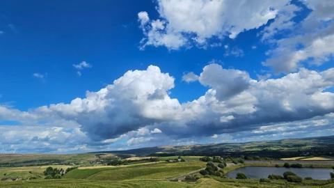 Blue sky is filled with large white and grey clouds with the sun glinting off the side of some. There are rolling hills and fields below with a large lake to the right.