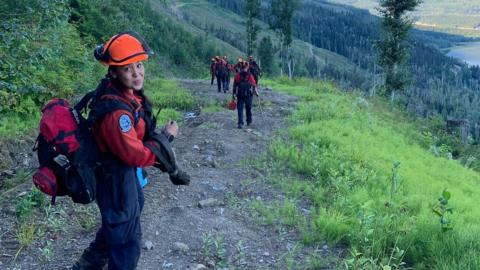 Devyn Gale, a young firefighter killed while tackling wildfires in Canada, is seen standing on a footpath on a mountainside in her firefighters' uniform. She is sticking out her tongue.