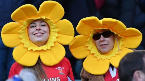 Wales supporters wearing daffodil hats smile in the crowd ahead of the Six Nations international rugby union match between England and Wales at Twickenham Stadium, west London, on February 26, 2022.