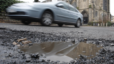 A car drives past a pothole filled with water in front of a church