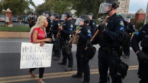 A woman with a 'Love Conquers All' sign confronts police officers with batons