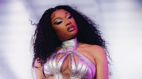 Megan Thee Stallion wearing a shiny outfit, looking to her left. The background is blurred.