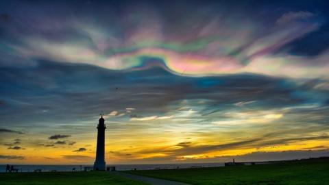 Rainbow coloured clouds wave across the top of the sky, with deep blue sky below. This is all over a slice of orange sky as the sun rises above a monument on the sea front in Sunderland