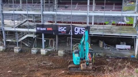 Blue/green digger picking up soil behind the partially-completed East Stand