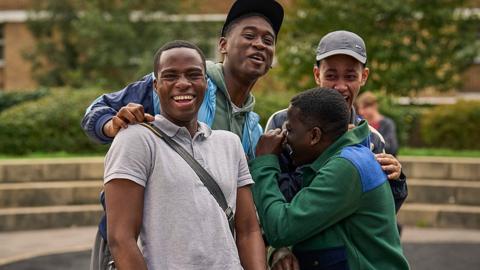 (L-R);Bayo (played by Juwon Adedokun, a young black man with short hear wearing a grey polo shirt who smiles at the camera), Bishop (played by Tienne Simon, a young black man wearing a black cap, green hoody and blue puffer jacket who smiles with his hand on Juwon's shoulder); Dane (played by Yus Jamal Crookes, a young black man with short hair wearing a green overshirt who holds his hand to his face which is turned slightly away from the camera as he laughs) and Junior (played by Gabriel Robinson, a young black man wearing a grey sports cap and dark jacket, standing slightly behind his castmates and laughing)