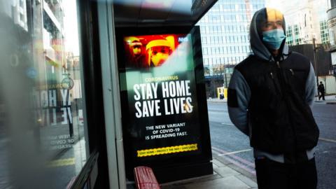 A man wears a mask at a bus stop with a Stay 91ȱ Save Lives poster