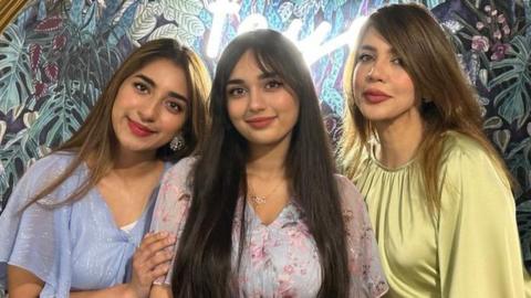 Aliza Ayaz (left) with her sister and mother. Aliza is a 25-year-old South Asian woman with long brown hair worn loose. She wears a sparkly blue blouse and leans into her sister, who also has long dark hair and wears a light pink top. Her mother is on the end and wears a pale green top. They are pictured in front of a wallpaper decorated with leaves and foliage.