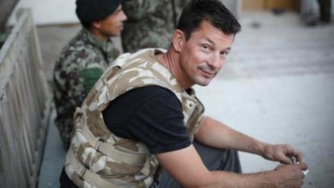Photojournalist John Cantlie in Syria. He is sat on a bench wearing a camouflage flak jacket and looking up at the camera.