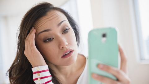 A woman looks at her smartphone while rubbing her temples as though suffering from a headache or stress