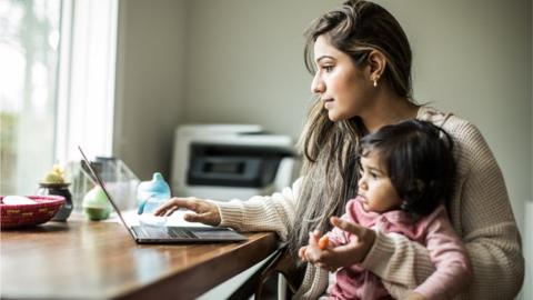 Woman holding a toddler while using a laptop.