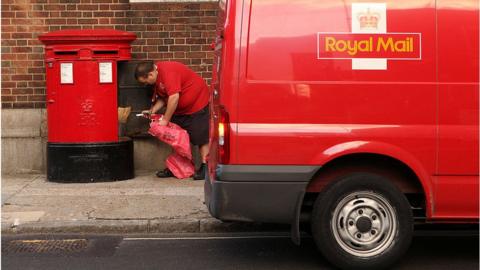 Royal Mail worker kneeling behind a Royal Mail red van getting post out of a red post box