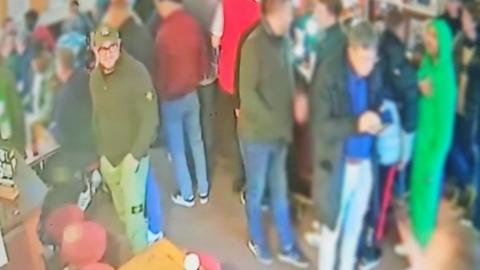 CCTV image of a man in green with a hat and glasses stood in a crowd of people at a pub