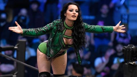 Saraya stands on the turnbuckle of the wrestling ring, wearing a green leather jacket. Her arms are outstretched as she interacts with the crowd