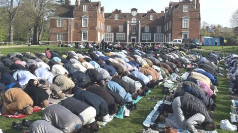 a large group of Muslim people bowing forward in prayer on the lawn in front of a Tudor mansion