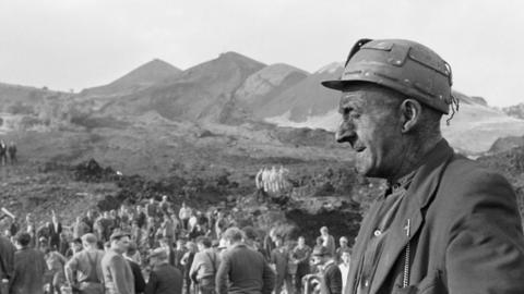 Miner with eyes closed wearing pit helmet at Aberfan disaster, as recovery continues behind him
