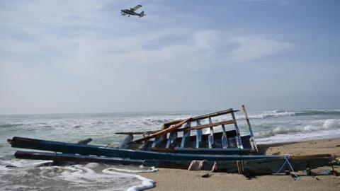 Broken pieces of a boat on a beach, as a plane flies overhead