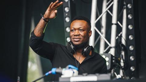 Benga looks out from behind a DJ desk on stage, his had raised