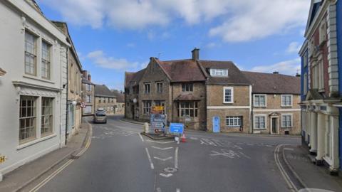 Google Maps view of the junction of the A359 High Street and Coombe Street in Bruton Town Centre. Two lanes can be seen, with numerous road signs at the junction line. There are buildings either side of the road and opposite the junction.
