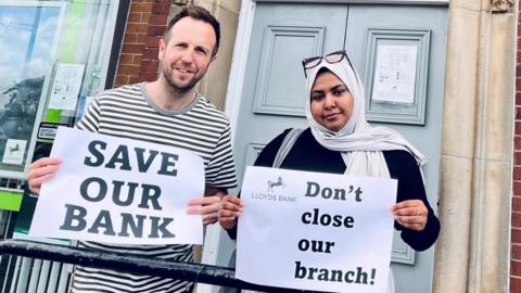 Councillors Ben Miskell and Nabeela Mowlana standing outside Lloyds Bank holding placards