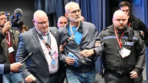 Andrew Boff being escorted out of the conference