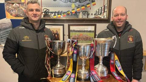 Two men flank a table of trophies
