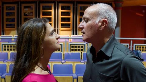 A woman and a man stare each other down, nose to nose, in a theatre rehearsal room