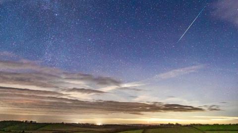 A meteor flies through a blue sky in the top right of the picture as the sun sets on the horizon