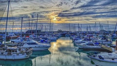 Boats in a harbour are shades of blue and white with the water reflecting the sunrise above. The sky if full of wispy lines of blue in a cream background